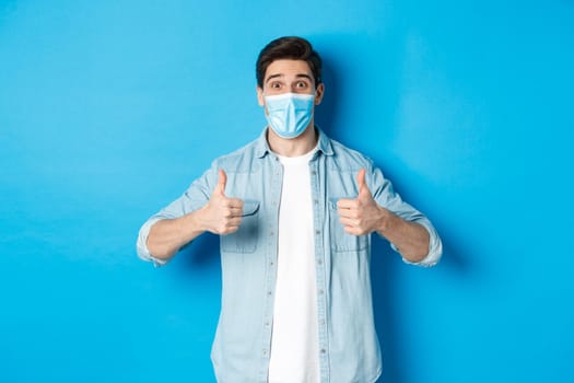 Concept of covid-19, pandemic and social distancing. Amazed guy in medical mask showing thumbs-up, recommending promo offer, standing against blue background.