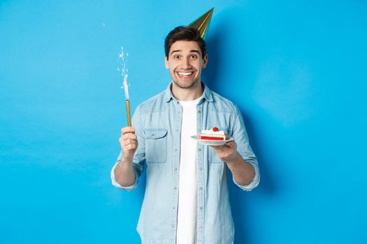 Happy young man celebrating birthday in party hat, holding b-day cake and smiling, standing over blue background.
