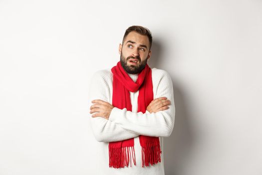 Confused man looking doubtful, standing with hands crossed and feeling cold on winter, wearing christmas sweater with red scarf, standing over white background.