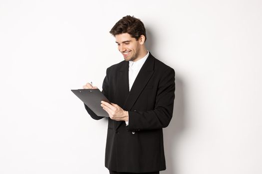 Image of handsome businessman in suit signing documents, looking at clipboard and smiling, standing against white background.