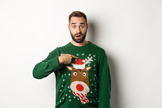 Winter holidays and christmas. Confused bearded guy pointing at himself, being startled with offer, standing over white background in sweater.