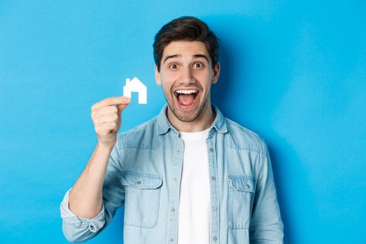 Real estate concept. Happy young man looking excited, found apartment, showing small house model, standing over blue background.