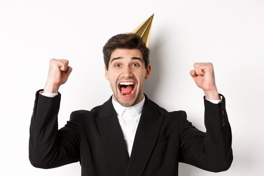 Close-up of happy good-looking man, wearing party hat and suit, raising hands up and rejoicing, celebrating new year, standing against white background.