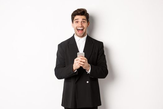 Image of excited handsome guy receiving promo offer on mobile phone, looking amazed and smiling, standing against white background.