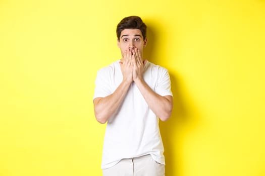 Handsome man looking shocked and speechless, holding hands on mouth, standing over yellow background.