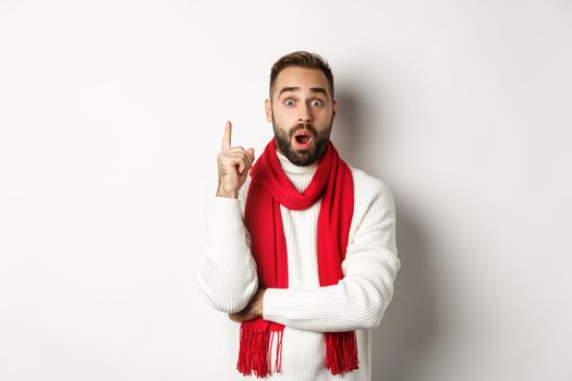 Christmas holidays and celebration concept. Excited bearded man having an idea, raising finger and suggesting plan, standing in red scarf with sweater, white background.
