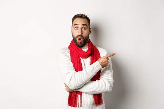 Christmas holidays and celebration concept. Surprised bearded man pointing finger right and looking impressed at camera, standing in red scarf and sweater, white background.