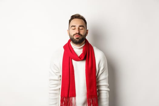Handsome man standing with closed eyes and puckered lips, waiting for christmas kiss under mistletoe, standing over white background in winter scarf and sweater.