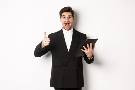 Portrait of confident businessman in black suit, holding clipboard with documents and showing thumb-up in approval, praise good job, standing against white background.