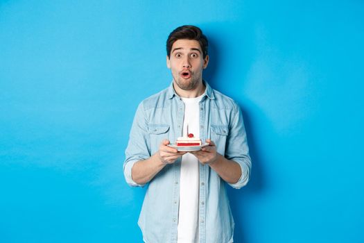 Man holding birthday cake and looking surprised, making a wish on lit candle, standing over blue background.