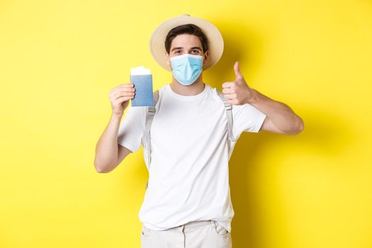 Concept of covid-19, tourism and pandemic. Happy male tourist in medical mask showing passport, going on vacation during coronavirus, make thumb up sign, yellow background.