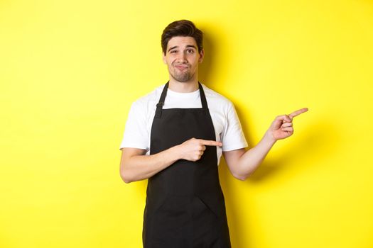 Doubtful barista in black apron pointing fingers right, looking skeptical and unamused, standing over yellow background.
