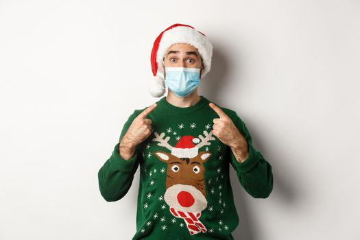 Christmas during pandemic, covid-19 concept. Man in in Santa hat pointing at his face mask, celebrating New Year and social distancing, white background.