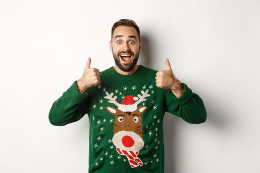 Christmas, holidays and celebration. Excited bearded guy in green sweater showing thumbs up, like something amazing, standing over white background.