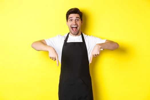 Waiter, coffee shop barista in black apron pointing fingers down, looking amazed, standing over yellow background.