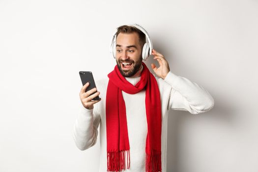 Winter holidays and technology concept. Happy man listening music in headphones, looking amazed at mobile screen, standing over white background.