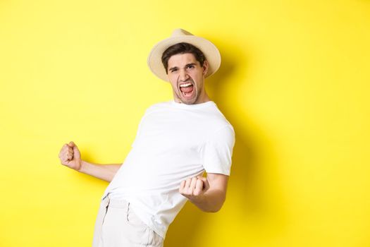 Concept of tourism and vacation. Satisfied young male tourist celebrating, winning something and rejoicing, making fist pump and shouting yes, standing against yellow background.