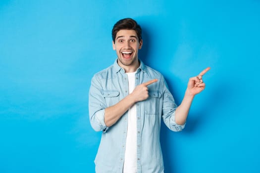 Image of excited handsome man in casual outfit, showing advertisement, pointing fingers right at copy space and smiling, standing against blue background.