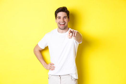 Confident smiling man pointing at you camera, standing in white clothes against yellow background.