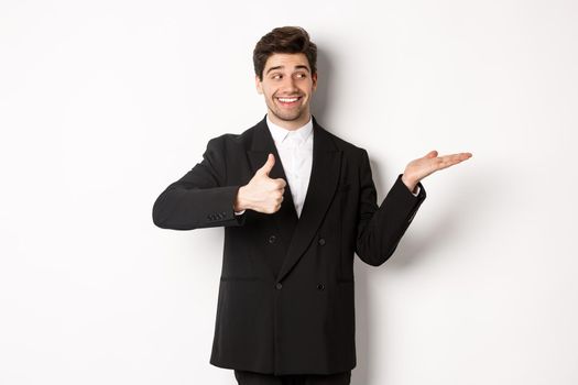 Handsome businessman in black suit, showing thumb-up and holding your product in hand over white copy space, standing against white background.