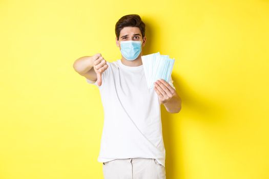 Concept of covid-19, quarantine and preventive measures. Man looking disappointed and showing thumb down, do not recommend bad medical masks, standing over yellow background.