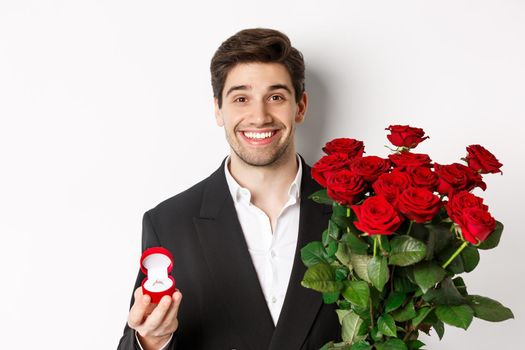 Close-up of attractive man in suit, holding bouquet of roses and engagement ring, making proposal, standing against white background.