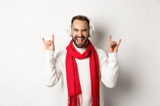 Joyful guy listening heavy metal in headphones, showing rock-on gesture and shouting for joy, standing over white background.