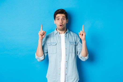 Portrait of amazed handsome man pointing fingers up, showing logo on blue background.