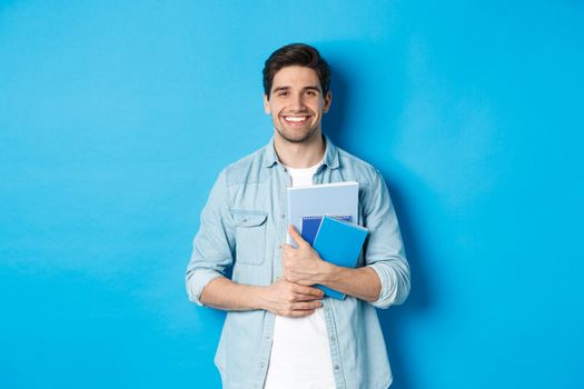 Young man holding notebooks and study material, smiling happy, standing over blue background.