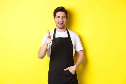 Handsome barista in black apron welcome guests to coffee shop, pointing finger gun and winking, standing against yellow background.