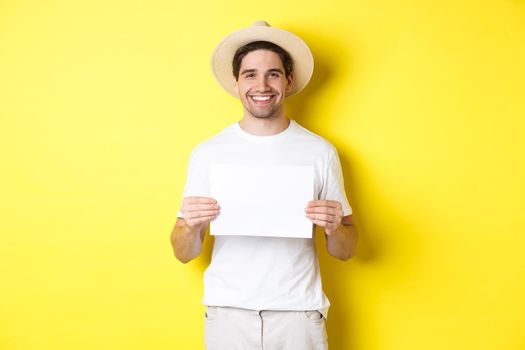 Handsome young male tourist in summer hat smiling, holding blank piece of paper for your sign, standing over yellow background.