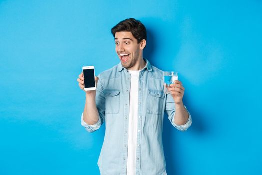 Happy man looking excited at mobile phone screen, holding glass of water, standing over blue background.