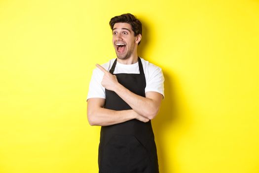 Waiter in black apron checking out promo offer, pointing finger and looking left at your logo, standing over yellow background.