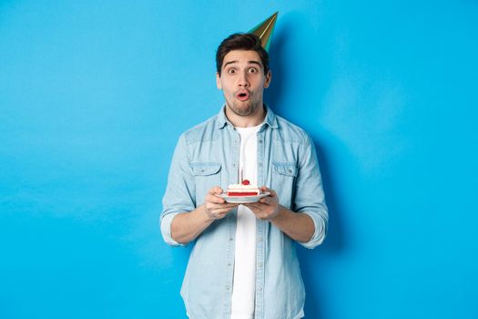 Handsome man in party cone holding birthday cake, looking surprised, standing over blue background.
