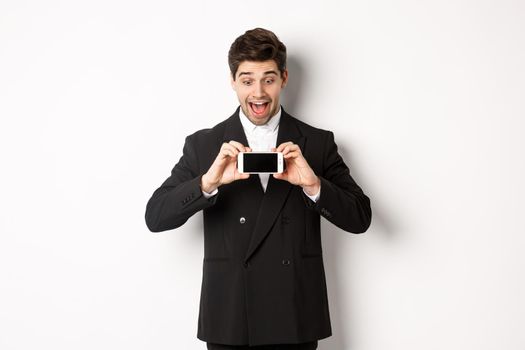 Portrait of excited handsome man showing smartphone screen, looking amazed at display with advertisement, standing over white background in black suit.