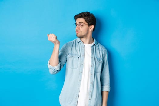 Young handsome man in glasses looking at his fingernails, checking manicure, standing over blue background.