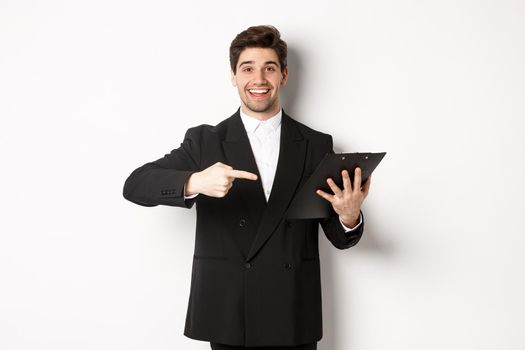 Image of handsome smiling businessman in black suit, pointing finger at clipboard with documents, standing against white background.