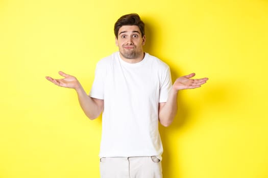 Clueless man shrugging, spread hands sideways confused, dont know anything, standing over yellow background.