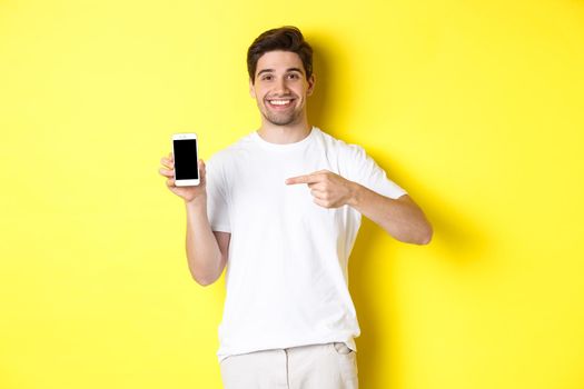 Image of attractive young man pointing finger at smartphone screen, showing an app, standing against yellow background.