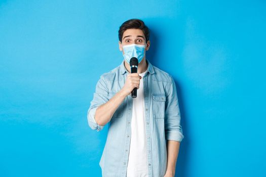 Young man in medical mask giving speech, holding microphone and looking confused, standing over blue background.