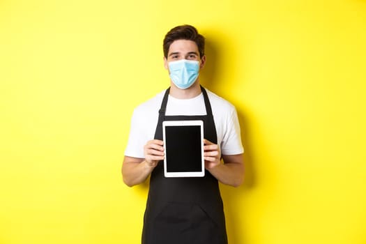 Concept of covid-19, small business and pandemic. Smiling barista in medical mask showing tablet screen, standing over yellow background.