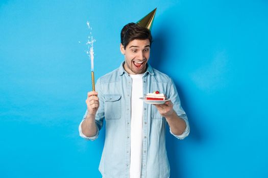 Happy young man celebrating birthday in party hat, holding b-day cake and smiling, standing over blue background.