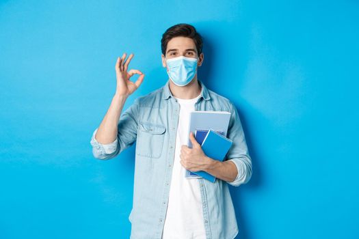 Education, covid-19 and social distancing. Guy student in medical mask looking happy, holding notebooks, showing ok sign, standing over blue background.
