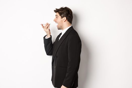 Profile shot of handsome businessman in black suit talking on speakerphone, smiling and looking happy, recording voice message, standing over white background.