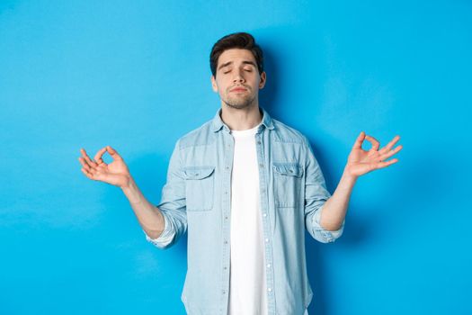 Calm man with closed eyes meditating, holding hands sideways and do yoga breathing exercises, standing against blue background.