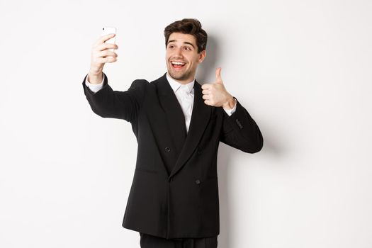 Portrait of good-looking man taking selfie on new year party, wearing suit, taking photo on smartphone and showing thumbs-up, standing against white background.