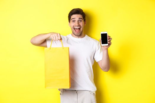 Concept of mobile banking and cashback. Young happy guy holding shopping bag and showing smartphone screen, yellow background.