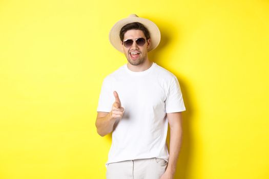 Confident and cheeky guy on vacation flirting with you, pointing finger at camera and winking, wearing summer hat with sunglasses, yellow background.