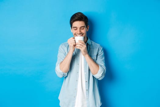 Young man drinking coffee with happy face, standing over blue background.