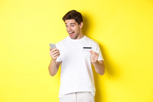 Man looking surprised at smartphone, shopping online, holding credit card, standing over yellow background. Copy space